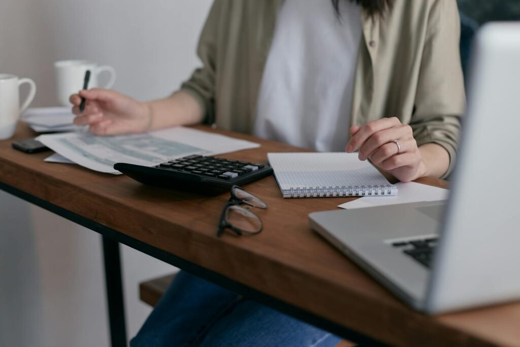 Contactar con Mostoles Asesores A woman manages finances at home, using a laptop and calculator on a wooden desk.